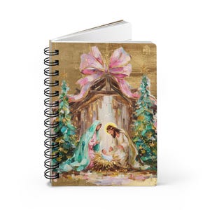 May include: A spiral-bound notebook featuring a gold-toned cover with a painted nativity scene. The artwork depicts a manger with figures, two decorated Christmas trees, and a large pink bow at the top, all rendered in a textured, painterly style.