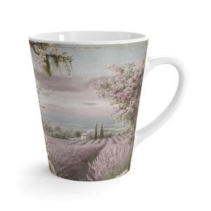 Może przedstawiać: Biały ceramiczny kubek do latte z sielskim krajobrazem pól lawendy, drzew i małego domu. Kubek ma zakrzywiony uchwyt i zwężający się kształt. Grafika zawiera tekst z boku kubka.