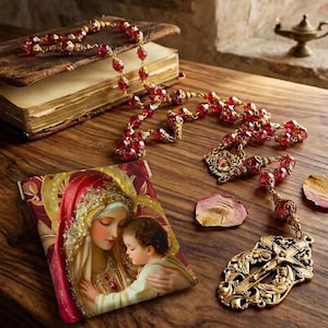 May include: A set consisting of a rectangular pouch featuring a printed image of a woman and child, paired with a rosary made of faceted red beads and a gold-toned metal crucifix. The rosary is draped across an old book with worn pages.