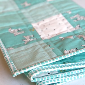 May include: A folded, quilted blanket with a teal and white color scheme. The blanket features a patchwork design with squares of teal fabric with white bunny illustrations and white squares with black dots. The edges are trimmed with a white and teal patterned border.