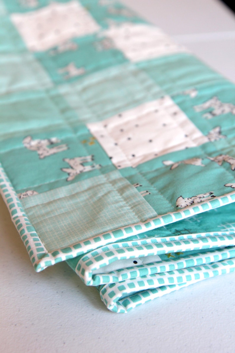 May include: A folded, quilted blanket with a teal and white color scheme. The blanket features a patchwork design with squares of teal fabric with white bunny illustrations and white squares with black dots. The edges are trimmed with a white and teal patterned border.