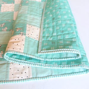 May include: A folded, handmade quilt featuring a patchwork design. The quilt showcases squares of white, light blue, and patterned fabric with dog illustrations. The edges are finished with a white and light blue border.