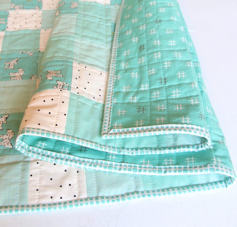 May include: A folded, handmade quilt featuring a patchwork design. The quilt showcases squares of white, light blue, and patterned fabric with dog illustrations. The edges are finished with a white and light blue border.