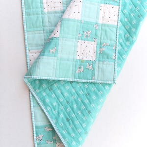 May include: A handmade baby quilt in shades of teal and white. The quilt features a patchwork design with squares of solid teal, white with black dots, and white with goat illustrations. The edges are finished with a white and teal trim.