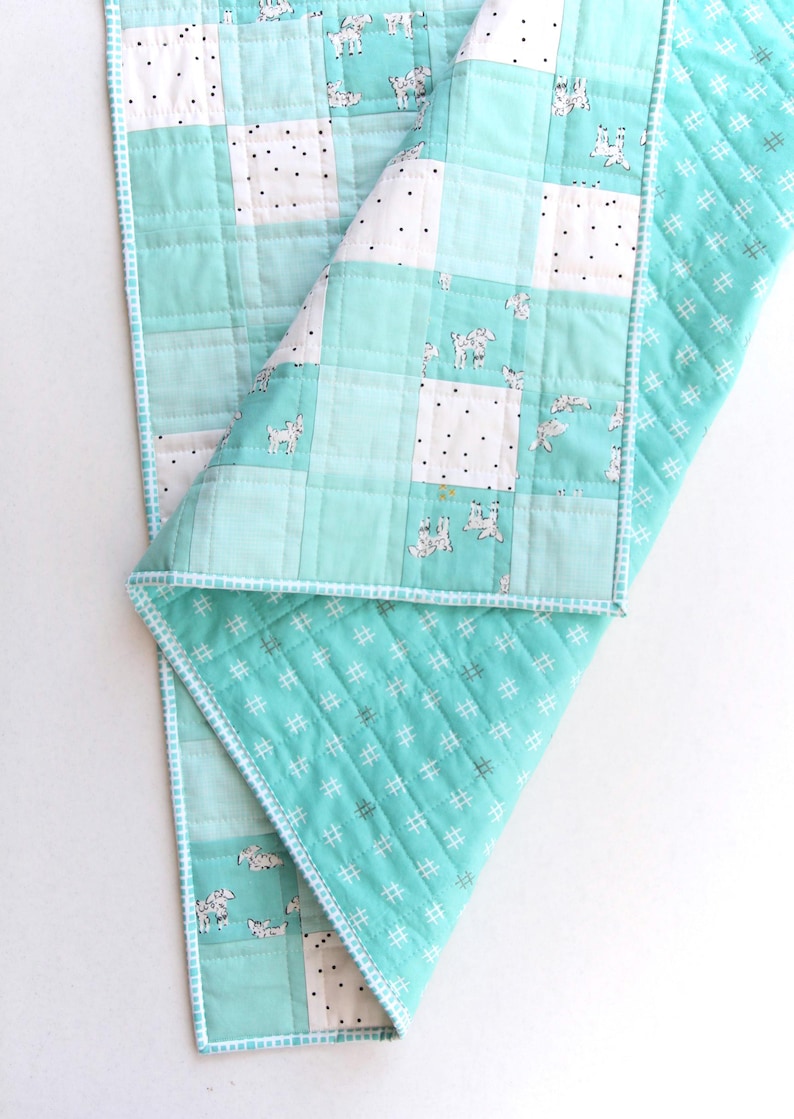 May include: A handmade baby quilt in shades of teal and white. The quilt features a patchwork design with squares of solid teal, white with black dots, and white with goat illustrations. The edges are finished with a white and teal trim.
