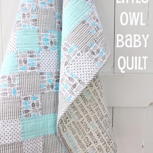 May include: A handmade baby quilt with a patchwork design. The quilt features squares of blue, gray, and white fabrics with owl and stripe patterns. The border has text. The words "Little Owl Baby Quilt" are visible.