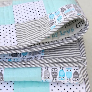May include: A folded patchwork quilt featuring a variety of patterns. The quilt incorporates squares of white with black polka dots, gray and white stripes, and solid light blue. The edges are trimmed with a gray and white striped fabric. The quilt has an owl pattern.