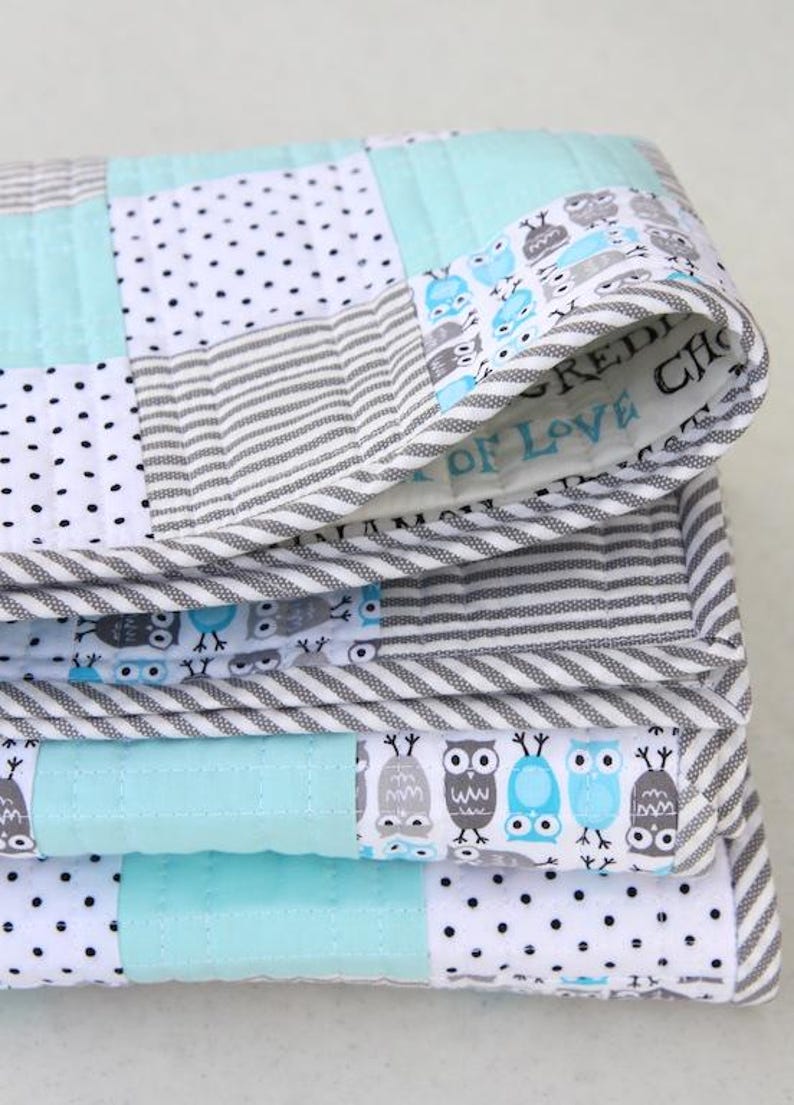 May include: A folded patchwork quilt featuring a variety of patterns. The quilt incorporates squares of white with black polka dots, gray and white stripes, and solid light blue. The edges are trimmed with a gray and white striped fabric. The quilt has an owl pattern.