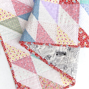 Può includere: Una trapunta patchwork con una varietà di tessuti fantasia in colori pastello. La trapunta presenta triangoli di tessuti rosa, blu, gialli e bianchi, con un bordo floreale rosso. Alcune sezioni hanno testo.