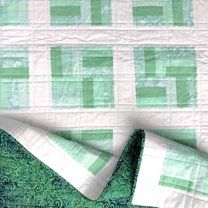 Op de afbeelding: Een handgemaakte quilt met een witte achtergrond en lichtgroene rechthoekige blokken. De onderkant van de quilt heeft een donkergroene stof met een wervelend patroon. De randen zijn afgewerkt met witte binding.