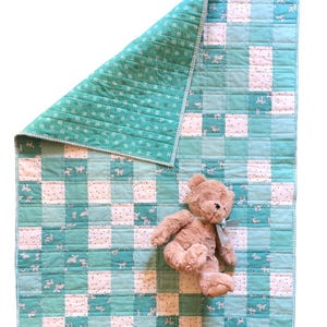 May include: A handmade baby quilt with a patchwork design in shades of teal and white, featuring a pattern of small animals. The quilt is paired with a tan teddy bear with a blue ribbon. The quilt measures approximately 40 x 40 inches.