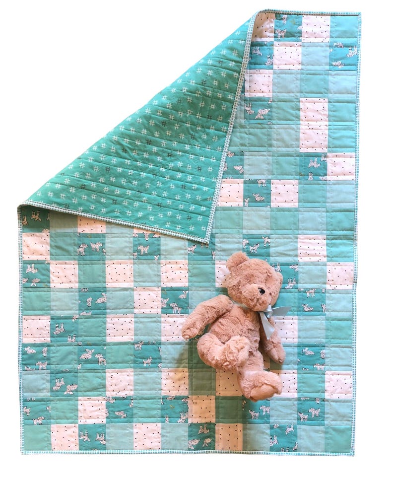 May include: A handmade baby quilt with a patchwork design in shades of teal and white, featuring a pattern of small animals. The quilt is paired with a tan teddy bear with a blue ribbon. The quilt measures approximately 40 x 40 inches.