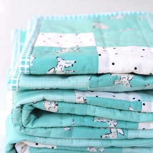 May include: A stack of folded quilts in shades of teal and white. The quilts feature a repeating pattern of sheep and black polka dots. The edges are finished with a contrasting border. The quilts appear soft and quilted.