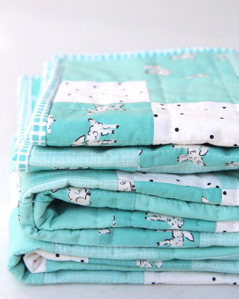 May include: A stack of folded quilts in shades of teal and white. The quilts feature a repeating pattern of sheep and black polka dots. The edges are finished with a contrasting border. The quilts appear soft and quilted.
