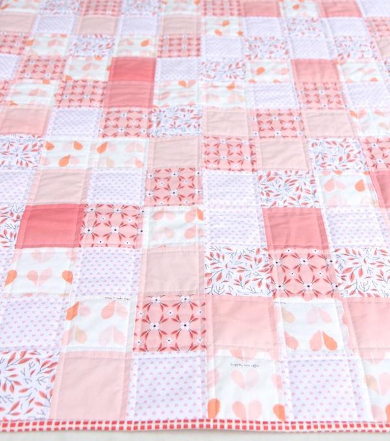 May include: A patchwork quilt featuring a variety of pink and white fabrics. The quilt is composed of numerous squares, some solid pink, others with floral or dotted patterns. The edges are finished with a red and white striped border.