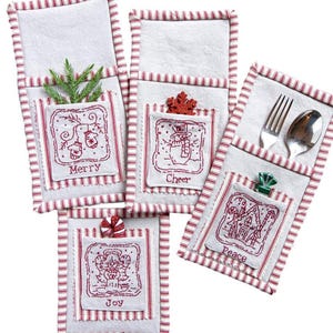 Puede incluir: Portacubiertos blancos con temática navideña con ribete a rayas rojas y bolsillos bordados. Cada bolsillo presenta un diseño festivo diferente y la palabra: Merry, Cheer, Joy y Peace. Un tenedor y una cuchara están en un soporte.