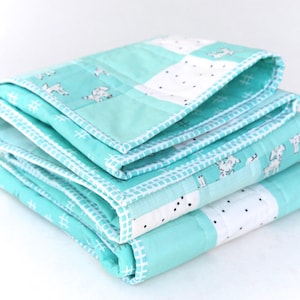 May include: A stack of three folded baby quilts. The quilts are a light turquoise color with white accents. The quilts have a patchwork design with a repeating pattern of white dogs and black dots. The edges are finished with a white stitched border.