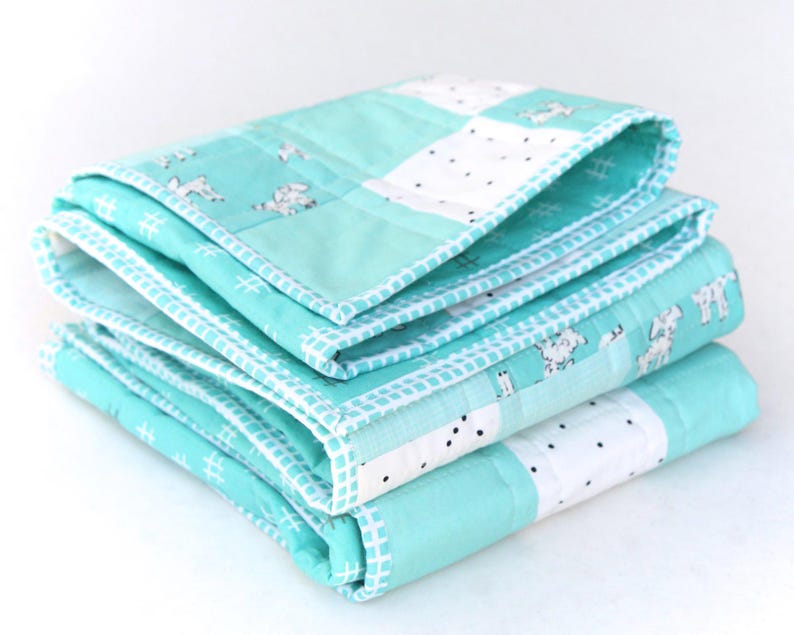 May include: A stack of three folded baby quilts. The quilts are a light turquoise color with white accents. The quilts have a patchwork design with a repeating pattern of white dogs and black dots. The edges are finished with a white stitched border.