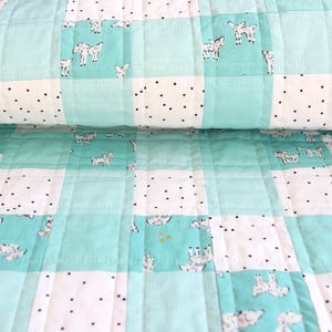 May include: A handmade quilt featuring a patchwork design with squares of white, light blue, and patterned fabric. The white squares have black polka dots, while the light blue squares have a goat pattern. The quilt is folded.