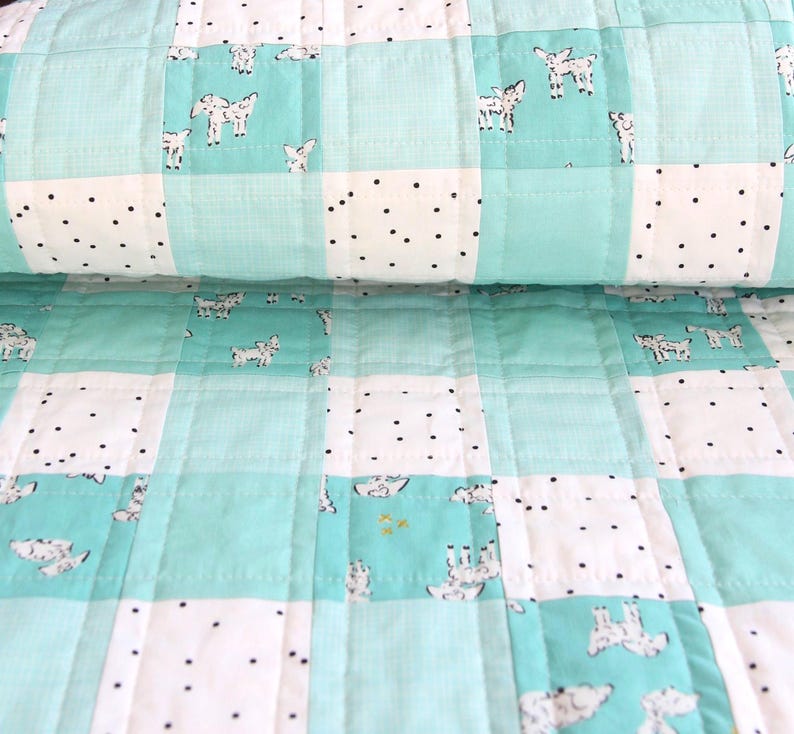 May include: A handmade quilt featuring a patchwork design with squares of white, light blue, and patterned fabric. The white squares have black polka dots, while the light blue squares have a goat pattern. The quilt is folded.