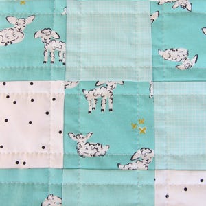 May include: A patchwork quilt featuring a repeating pattern of white sheep on a teal background. The quilt is constructed from squares of teal, white with black dots, and light teal fabric, all stitched together with white thread.
