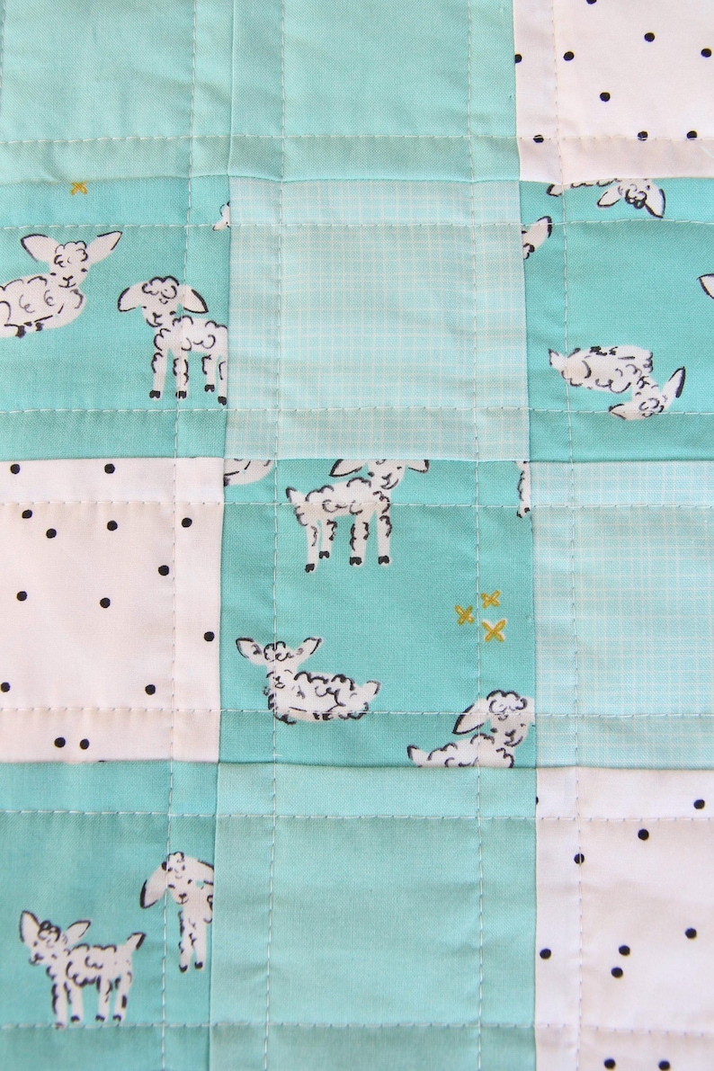 May include: A patchwork quilt featuring a repeating pattern of white sheep on a teal background. The quilt is constructed from squares of teal, white with black dots, and light teal fabric, all stitched together with white thread.