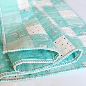 May include: A folded, quilted blanket in shades of teal and white. The blanket features a patchwork design with squares of solid teal, white with black polka dots, and white with a repeating animal print. The edges are trimmed with a white border.