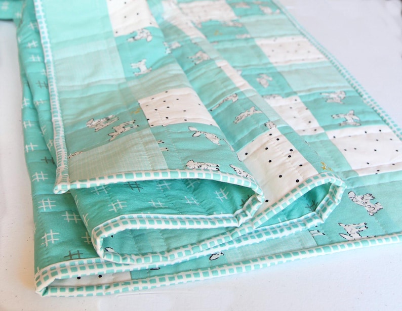 May include: A folded, quilted blanket in shades of teal and white. The blanket features a patchwork design with squares of solid teal, white with black polka dots, and white with a repeating animal print. The edges are trimmed with a white border.