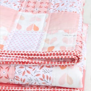 May include: Folded patchwork quilt in shades of pink, white, and coral. The quilt features floral and geometric patterns, with a red and white checkered border. The text "happily ever after" is visible on one of the quilt squares.