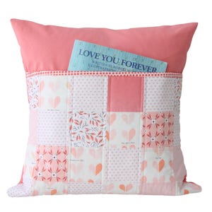 May include: A square, coral-colored pillow with a patchwork design on the front. The patchwork features various patterns of hearts and floral designs in shades of pink and white. A book with the title "LOVE YOU FOREVER" is tucked into a pocket on the pillow.