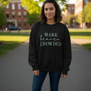 Puede incluir: Sudadera negra con el texto "MAKE heaven CROWDED" en escritura verde claro. La sudadera se usa con vaqueros azules y zapatillas blancas. La imagen está tomada al aire libre en un camino pavimentado.