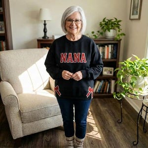 May include: A woman wearing a black sweatshirt with the word "NANA" in red block letters. The sweatshirt has red bow accents on the sides. She is wearing blue jeans and glasses, standing in a living room with bookshelves and plants.