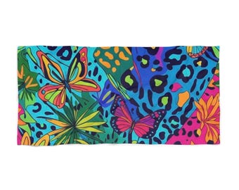 Puede incluir: Una toalla de playa rectangular con un diseño vibrante y colorido. El patrón presenta mariposas, flores y manchas de estampado de leopardo en tonos de azul, rosa, naranja y verde. La toalla está hecha de un material suave.