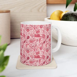 May include: White ceramic mug featuring a repeating pattern of foxes, owls, and floral elements in shades of pink and red. The mug has a curved handle and is sitting on a beige coaster. The design is whimsical and nature-themed.