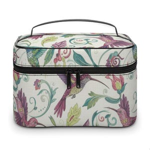 Può includere: Un beauty case rettangolare con cerniera e manico neri. La borsa presenta un motivo ripetuto di colibrì, fiori e foglie nei toni del viola, verde e giallo su sfondo crema. Ideale per i viaggi.