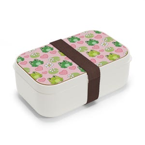 Puede incluir: Una fiambrera rectangular blanca con tapa rosa estampada con ranas verdes, corazones rosas y flores blancas. Una banda elástica marrón asegura la tapa. Ideal para transportar alimentos.