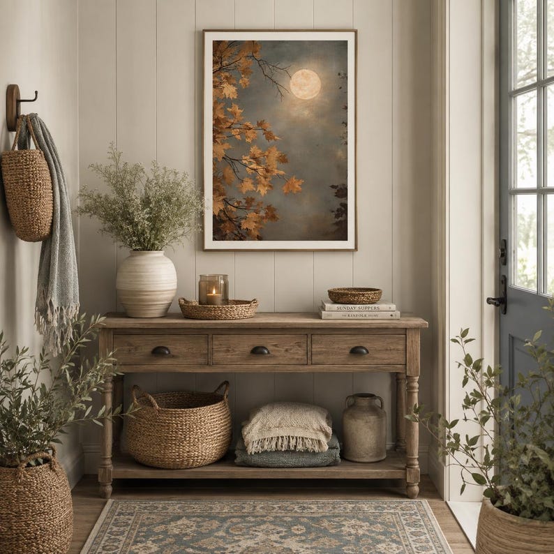 May include: A rectangular framed wall art print depicting a full moon over autumn maple branches with golden leaves. The artwork is displayed above a wooden console table featuring three drawers, a lower shelf, woven baskets, and a ceramic vase.
