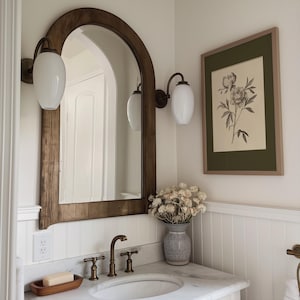 以下が含まれることがあります： 大きなアーチ型の木製鏡と2つの壁掛けライトを備えたバスルームインテリア。額入りの植物画が壁に掛かっています。大理石のカウンタートップには、洗面台、蛇口、ドライフラワーの花瓶があります。