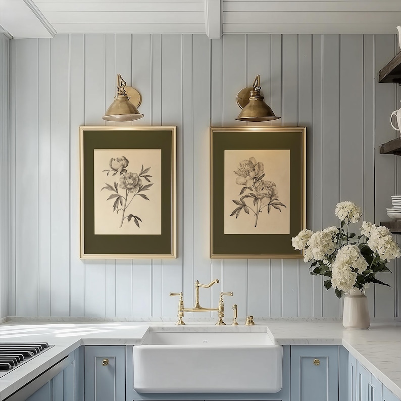Two vintage botanical peony art prints in gold frames with olive green mats displayed above a white farmhouse sink in a light blue paneled kitchen, styled with brass wall sconces, brass faucet, marble countertops, and a vase of white hydrangeas.