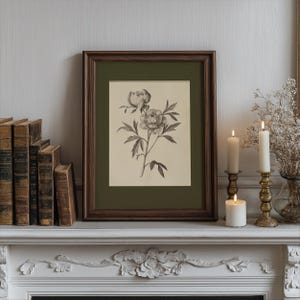 以下が含まれることがあります： 暗い木製のフレームに入ったシャクヤクの花の額入り植物画。このプリントは、白いマントルピースに、アンティークの本、火の灯ったろうそく、装飾的な真鍮の燭台とともに展示されています。全体的な美学はヴィンテージでエレガントです。
