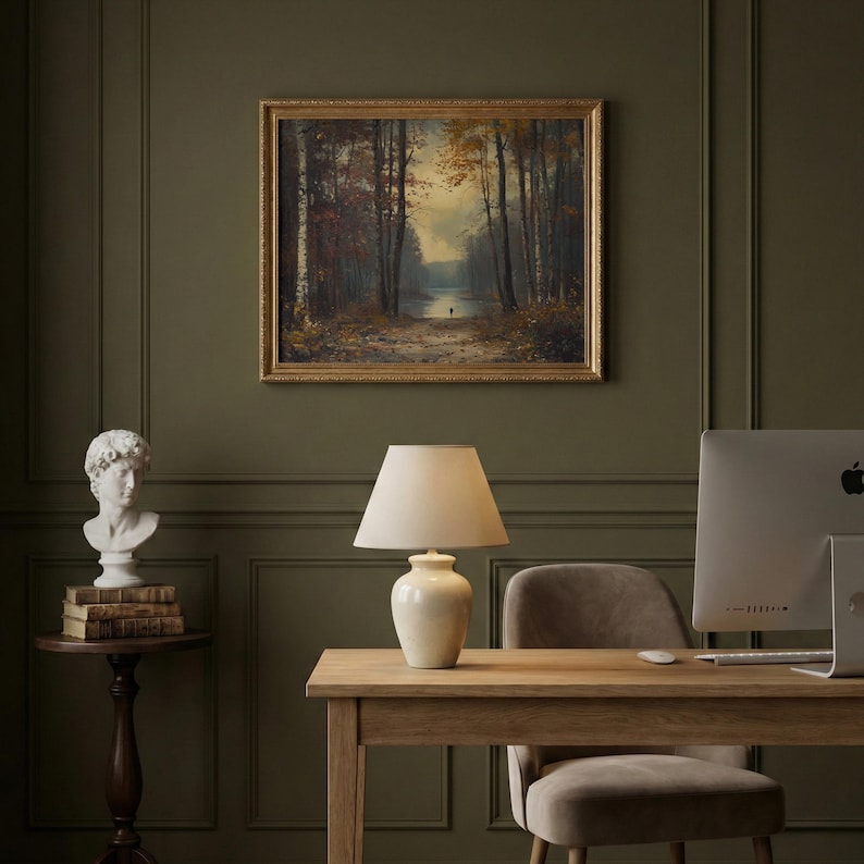 May include: A framed landscape painting depicting a forest path leading to a lake under a soft sky, displayed above a desk with a cream-colored ceramic table lamp, a white computer monitor, a gray chair, and a white bust on a small wooden table.