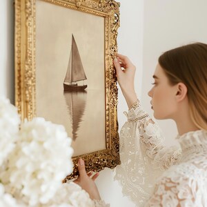 May include: A framed sepia-toned painting of a sailboat on calm water. The artwork is in an ornate, gold-colored frame. The image is being held up against a white wall.