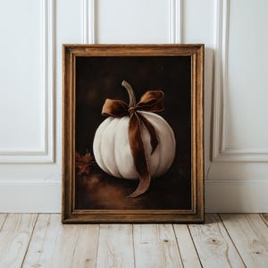 May include: A framed wall art print featuring a white pumpkin centered against a dark, moody background. The pumpkin is adorned with a long, flowing brown velvet ribbon tied in a bow around its stem. A single dried maple leaf sits to the left.
