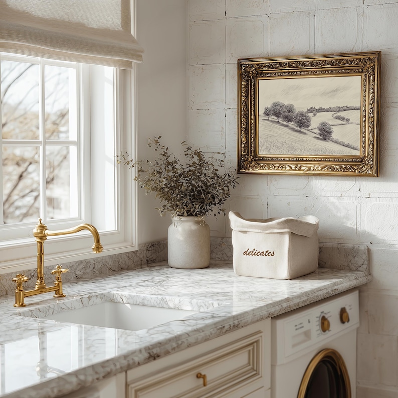 May include: A cream-colored fabric storage bin with the word "delicates" embroidered in brown thread on the front. The bin is positioned on a white marble countertop next to a ceramic vase filled with dried greenery and a gold faucet.