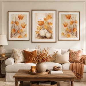 Könnte beinhalten: Ein Triptychon aus gerahmten Kunstdrucken mit Aquarellillustrationen von orangefarbenen und gelben herbstlichen Ahornblättern und zwei weißen Kürbissen. Die Kunstwerke sind in dünnen Goldrahmen über einem beigen Sofa angebracht.