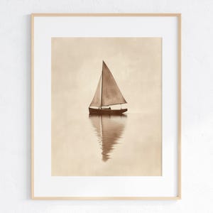 May include: A sepia-toned framed print of a sailboat on calm water. The sailboat has a triangular sail and is reflected in the water below. The artwork is in a light wooden frame, suitable for nautical or minimalist decor.
