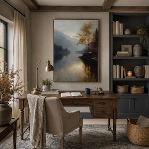 May include: A rectangular framed landscape painting depicting a calm lake surrounded by misty mountains and a golden-leafed tree, mounted on a wall above a wooden desk with a brass lamp, an open book, a dark mug, and a chair with a beige throw blanket.