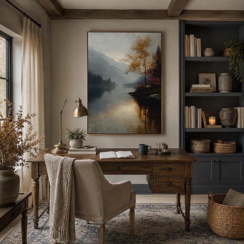 May include: A rectangular framed landscape painting depicting a calm lake surrounded by misty mountains and a golden-leafed tree, mounted on a wall above a wooden desk with a brass lamp, an open book, a dark mug, and a chair with a beige throw blanket.