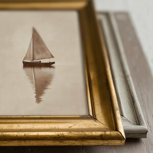 May include: A framed print of a sailboat on calm water. The artwork is in a gold frame, with a second, light-colored frame visible in the background. The sailboat is brown with a beige sail, and its reflection is visible in the water.
