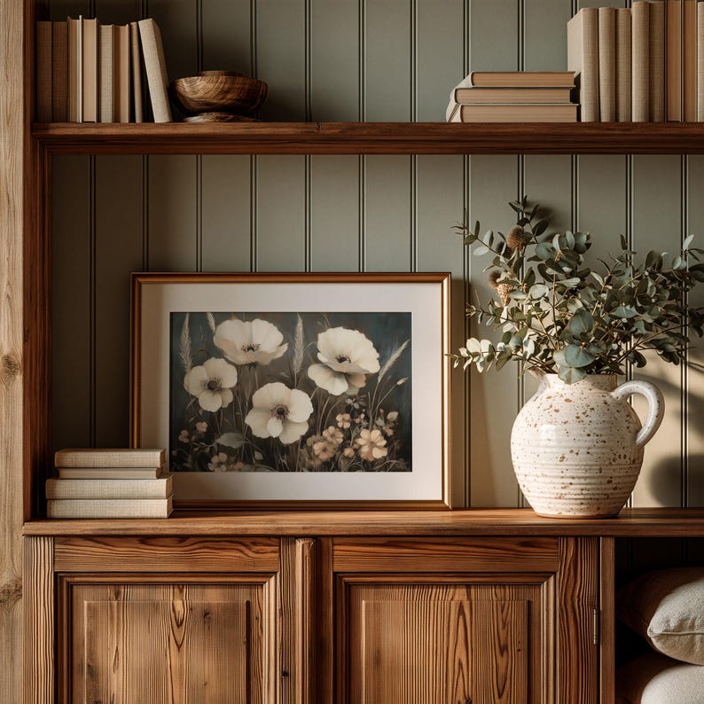 May include: A framed horizontal print featuring white poppy flowers and wheat stalks against a dark background, displayed on a wooden shelf next to a speckled cream ceramic pitcher filled with eucalyptus branches and a stack of books.
